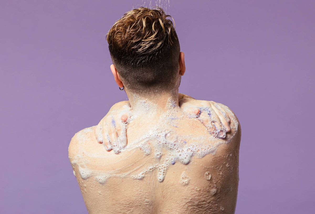Seen from behind, a person with short, damp hair washes in the shower, lathering Lush "Sleepy" shower gel over their shoulders and upper back. A subtle hint of purple pigment peeks through the white foam against a solid pastel lilac background.